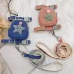 On a white surface, there are two small pet harnesses from the Dog Traction Rope Adjustable Pet Knapsack set. One harness is blue with a star patch labeled "Love," while the other is pink with a similar design. Each comes with its own matching leash—one blue and one pink—neatly coiled beside them. Beside the harnesses are glasses and flowers, adding to the scene.